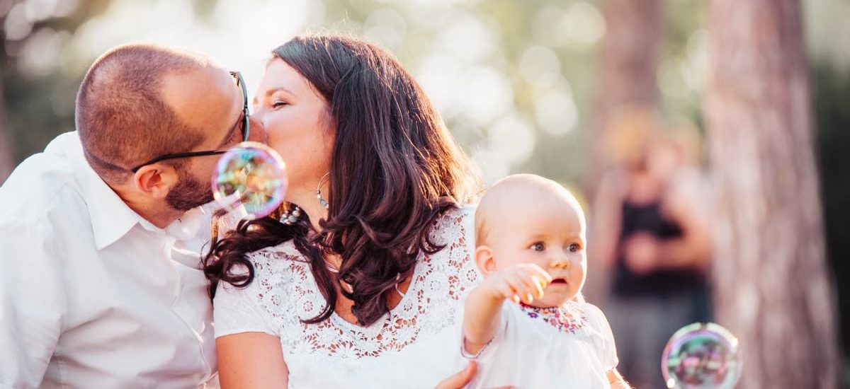 A tender family moment captured with a couple kissing and a baby holding bubbles outdoors.