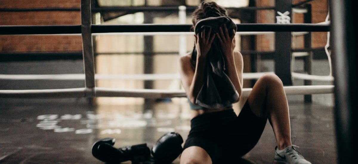 A female boxer sitting in a gym ring, resting and wiping sweat with a towel after a workout.